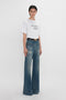 A person stands against a white background, donning a white T-shirt with the text "DAVID'S WIFE" and wide-legged Alina High Waisted Jean In Indigrey Wash by Victoria Beckham featuring seventies-style pockets.