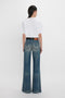 A person with dark hair stands facing away, wearing a white T-shirt, Victoria Beckham Alina High Waisted Jean In Indigrey Wash with seventies-style pockets, and a dark belt.