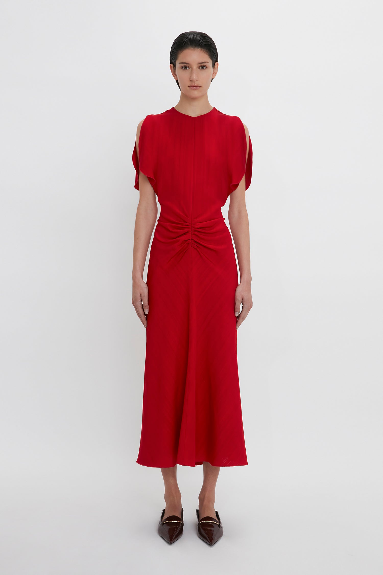 A person stands against a plain background, wearing an Exclusive Gathered Waist Midi Dress In Carmine by Victoria Beckham with open shoulders, along with pointy brown shoes.
