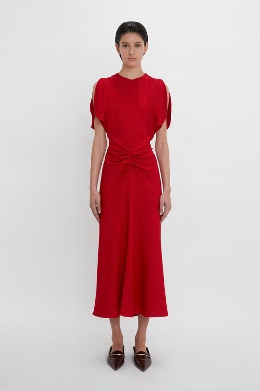 A person stands against a plain background, wearing an Exclusive Gathered Waist Midi Dress In Carmine by Victoria Beckham with open shoulders, along with pointy brown shoes.