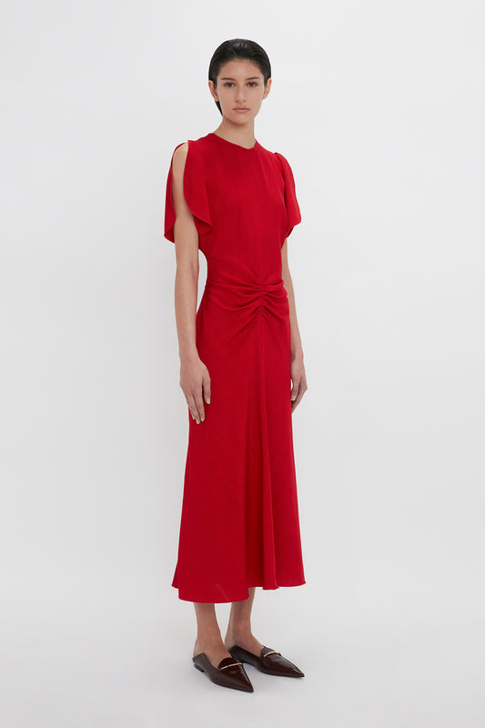 Person stands against a plain white background wearing the Victoria Beckham Exclusive Gathered Waist Midi Dress In Carmine. They have short dark hair and are wearing brown flat shoes, exuding a chic vibe reminiscent of Victoria Beckham's style.