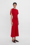 Person stands against a plain white background wearing the Victoria Beckham Exclusive Gathered Waist Midi Dress In Carmine. They have short dark hair and are wearing brown flat shoes, exuding a chic vibe reminiscent of Victoria Beckham's style.