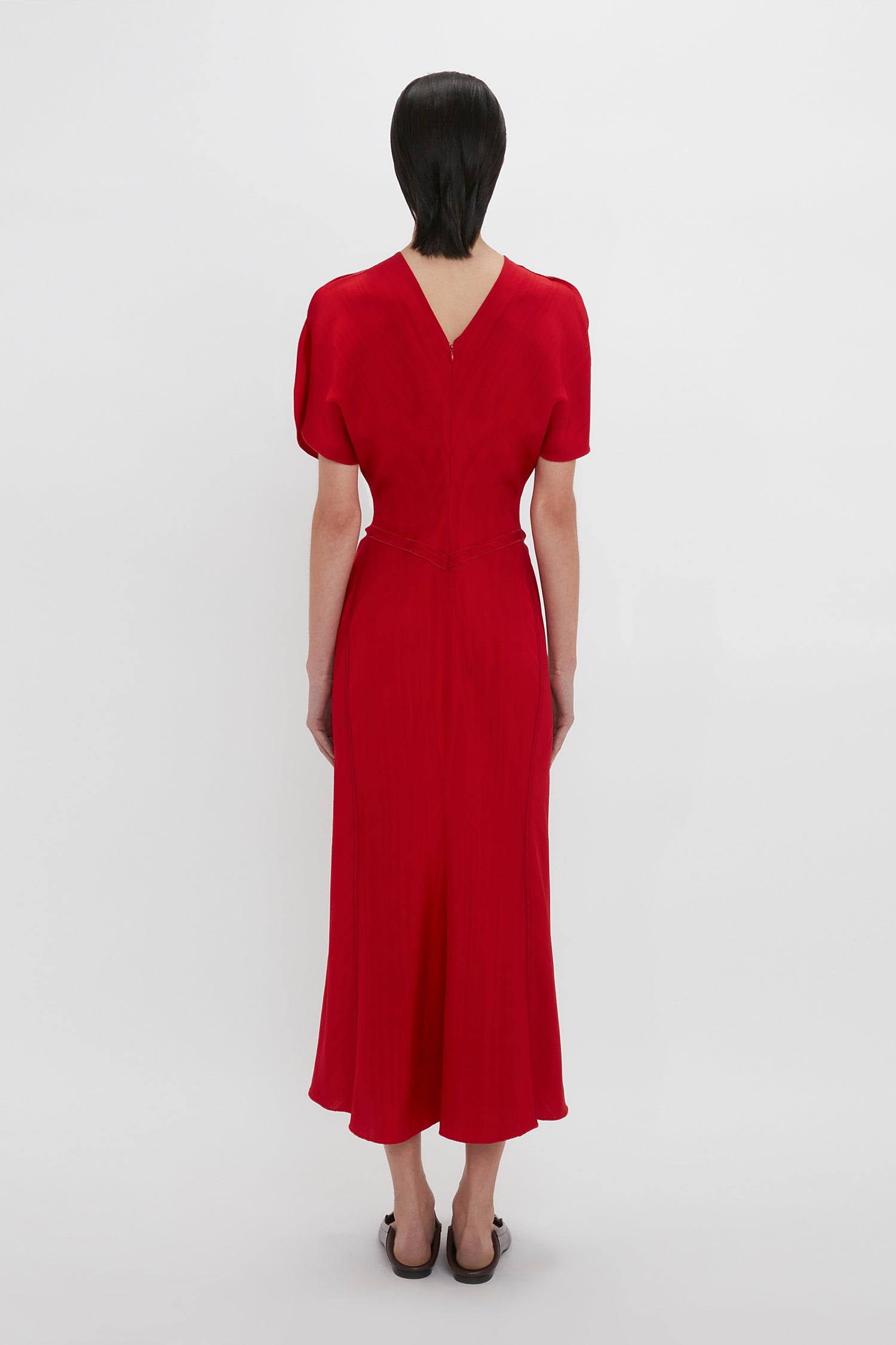A person with short, dark hair stands with their back to the camera, wearing a red Exclusive Gathered Waist Midi Dress In Carmine by Victoria Beckham, with short sleeves and a V-shaped back—channelling elegant Victoria Beckham vibes.