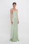 A person is standing and wearing a long, light green satin dress with thin straps against a plain white background, adorned with an Exclusive Camellia Flower Necklace In Gold handcrafted in Italy from gold brushed brass by Victoria Beckham.