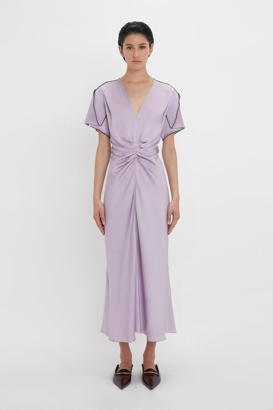 A person stands facing forward, wearing the Gathered V-Neck Midi Dress In Petunia from Victoria Beckham, an elegant light purple midi dress with a waist-defining pleat detail and short sleeves, paired with brown pointed shoes. The figure-flattering stretch fabric enhances the look against the plain white background.