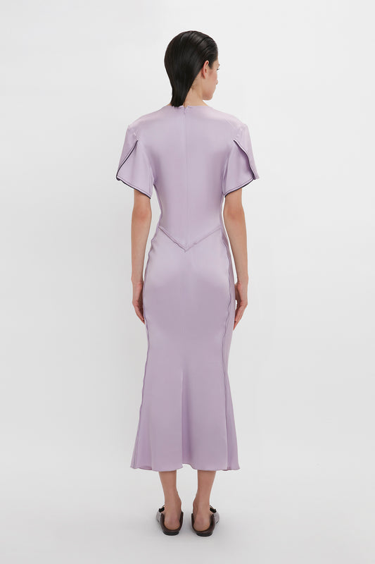 A person with short hair is seen from behind wearing a pale purple ankle-length dress with a high neckline, short sleeves, and a fitted bodice. The elegant Gathered V-Neck Midi Dress in Petunia by Victoria Beckham highlights the waist-defining pleat detail, made from figure-flattering stretch fabric for added comfort and style.