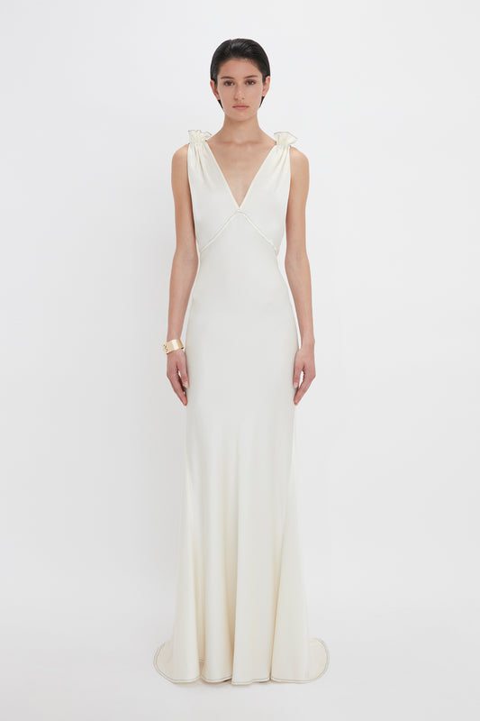 A person stands facing forward wearing a Victoria Beckham Gathered Shoulder Floor-Length Cami Gown In Ivory made of crepe back satin with a V-neckline and feminine frills. They have short, dark hair and are posing against a plain white background.