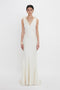 A person stands facing forward wearing a Victoria Beckham Gathered Shoulder Floor-Length Cami Gown In Ivory made of crepe back satin with a V-neckline and feminine frills. They have short, dark hair and are posing against a plain white background.