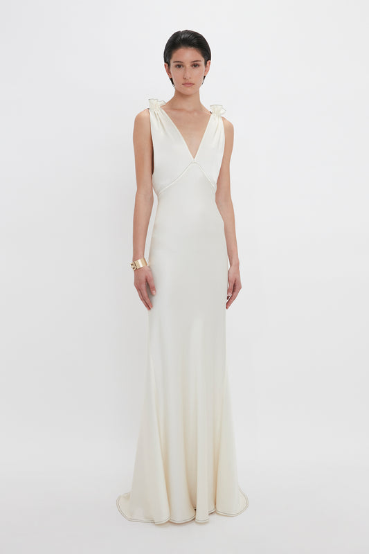 A person stands against a plain white background wearing a Gathered Shoulder Floor-Length Cami Gown in Ivory by Victoria Beckham. They also have a gold bracelet on their right wrist.