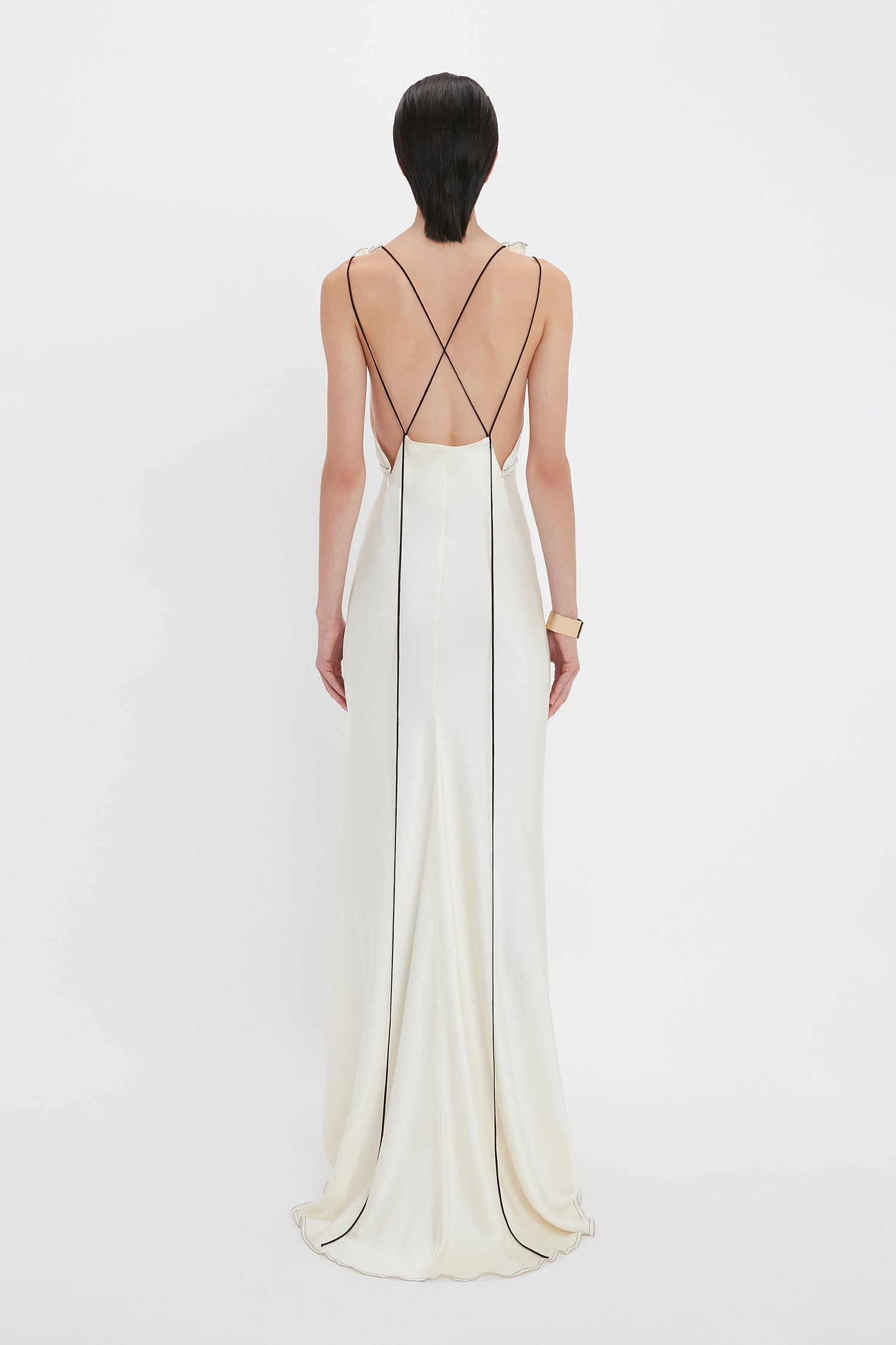 Person in a Victoria Beckham Gathered Shoulder Floor-Length Cami Gown in Ivory with thin black straps crisscrossing the back, standing against a plain white background.