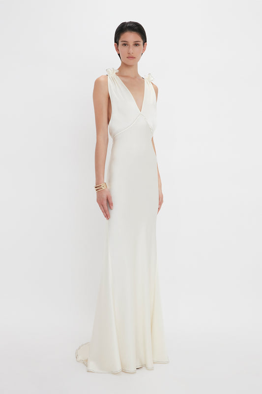 A woman stands against a plain white background wearing a sleeveless, floor-length Gathered Shoulder Floor-Length Cami Gown In Ivory by Victoria Beckham with a V-neck and slight train. Made of crepe back satin, the dress highlights her short dark hair and is accessorized with gold bracelets.
