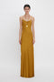 A person stands wearing a Victoria Beckham Low Back Cami Floor-Length Dress In Harvest Gold. They have short, dark hair and are adorned with a long necklace. The background is plain white.