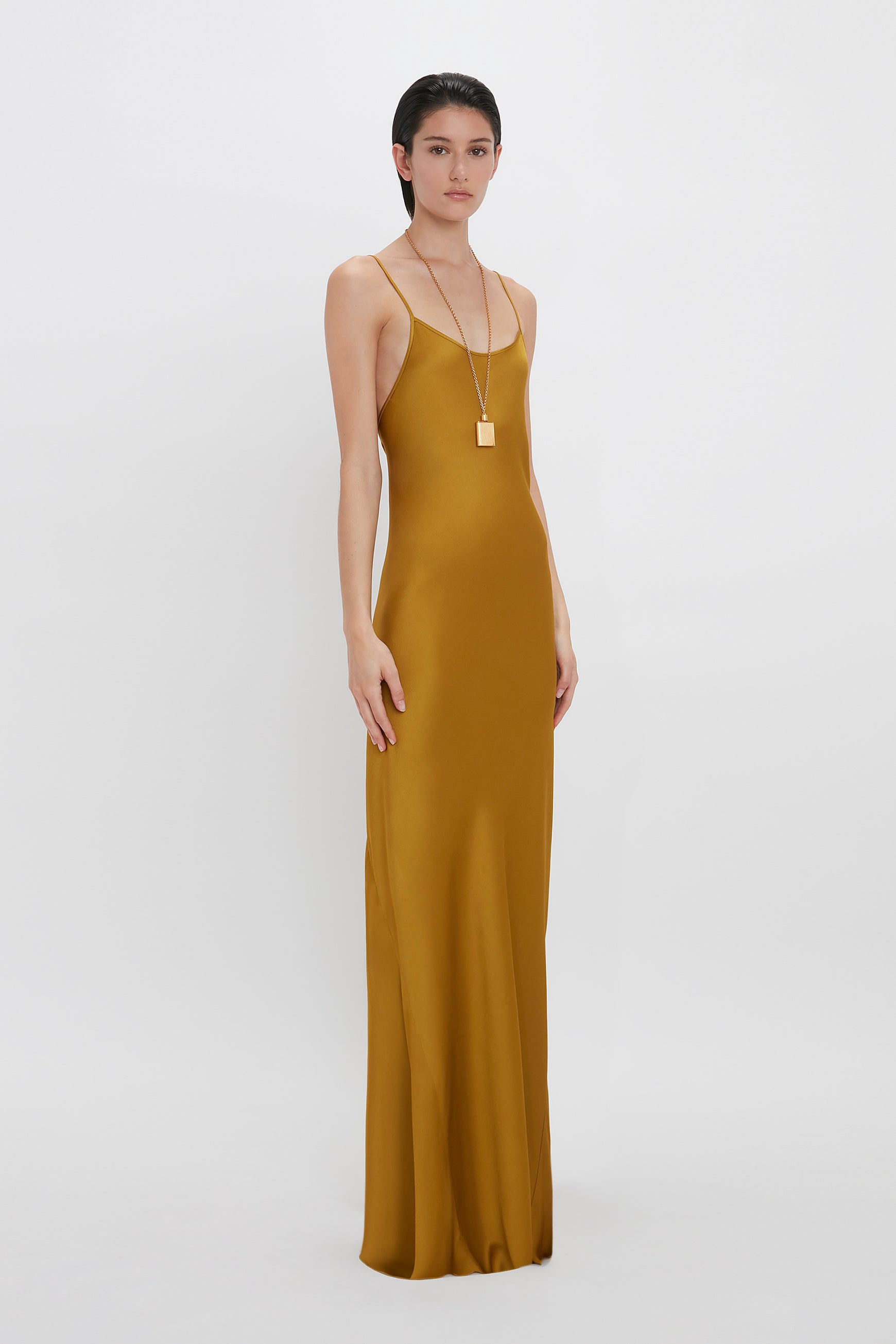 A person stands against a white background wearing a floor-length, Low Back Cami Floor-Length Dress In Harvest Gold by Victoria Beckham with spaghetti straps and a long necklace.