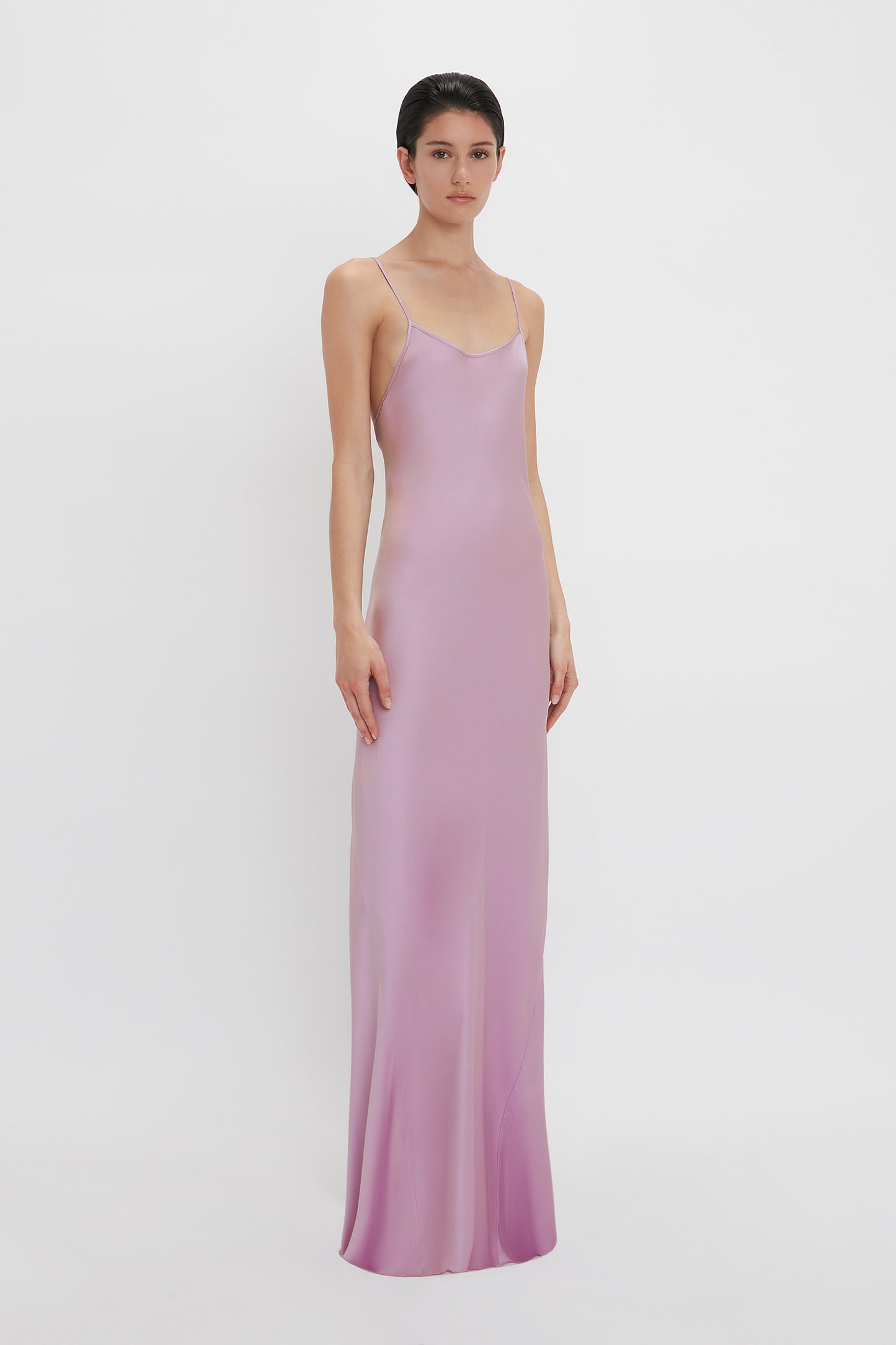 A person is standing against a plain white background, wearing the Victoria Beckham Low Back Cami Floor-Length Dress In Rosa, with thin straps that accentuate a sensuous silhouette.