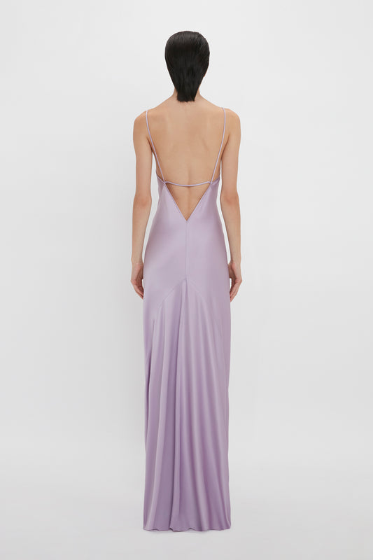 Rear view of a person wearing the Victoria Beckham Low Back Cami Floor-Length Dress In Petunia with spaghetti strap detailing, reminiscent of 1990s style, standing against a plain white background.