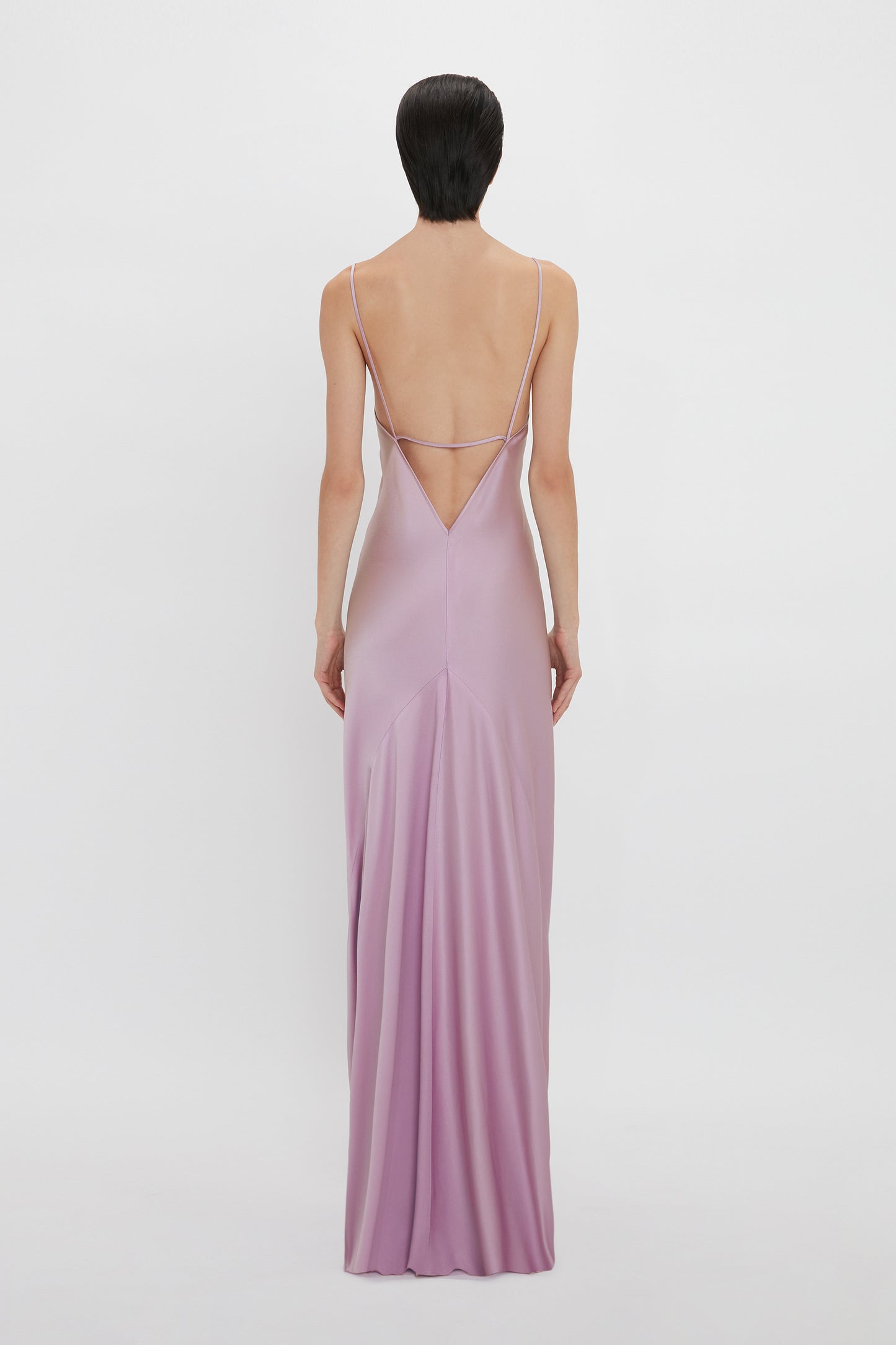 A person with short dark hair is wearing a Low Back Cami Floor-Length Dress In Rosa by Victoria Beckham, standing with their back to the camera, showcasing a sensuous silhouette.