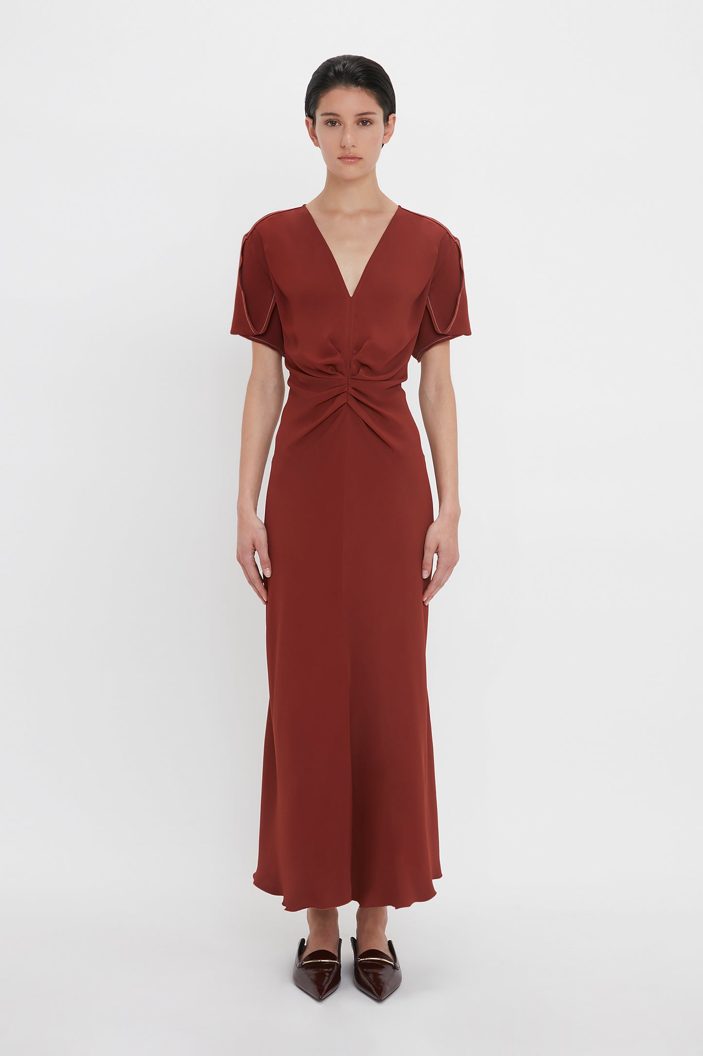 A person stands against a plain white background, wearing the Victoria Beckham Gathered V-Neck Midi Dress In Russet. The figure-flattering stretch fabric accents their shape perfectly. They complete the look with brown pointed shoes and short, dark hair.