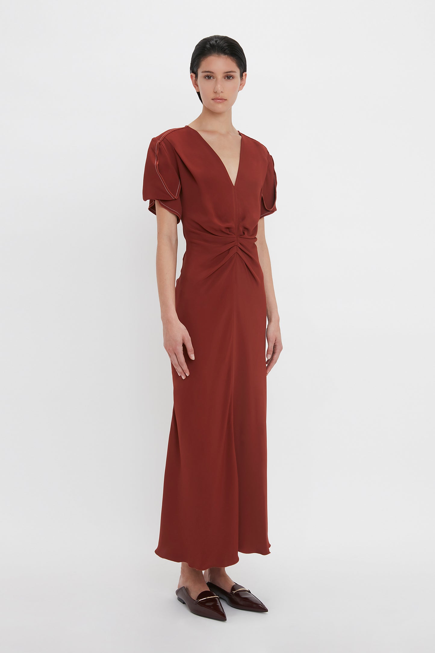 A person stands against a white background wearing a figure-flattering, deep red Gathered V-Neck Midi Dress In Russet by Victoria Beckham with short sleeves and brown pointed shoes. The dress features a waist-defining pleat detail that enhances the silhouette beautifully.