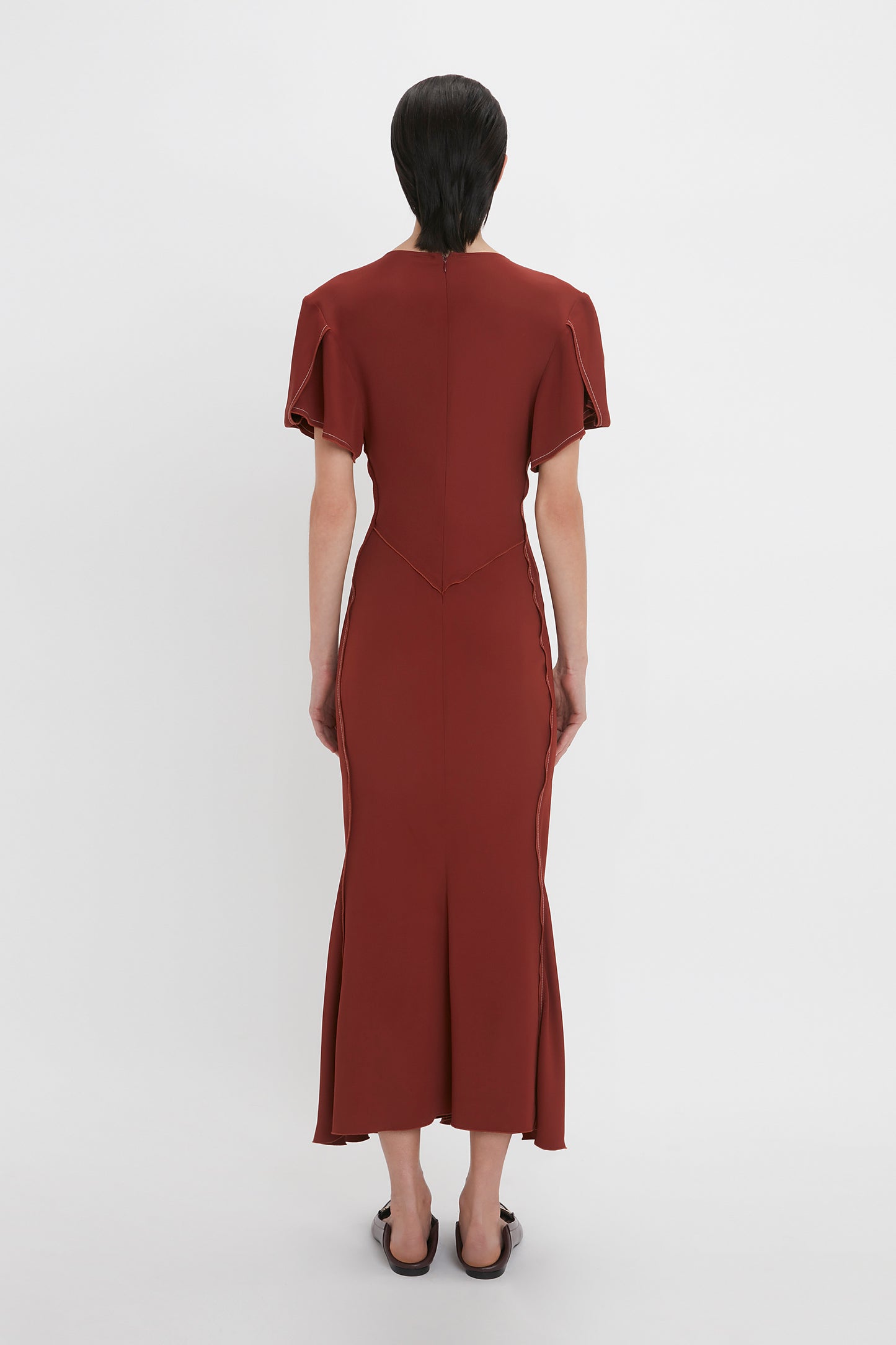 A person with short dark hair is standing facing away from the camera, wearing a Victoria Beckham Gathered V-Neck Midi Dress In Russet and dark flat shoes.