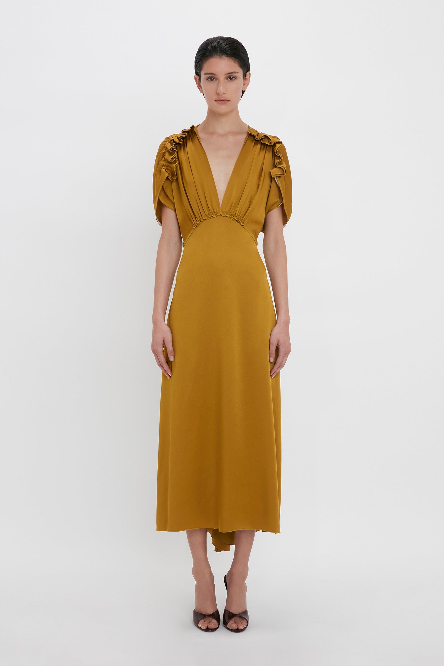 Woman wearing a Victoria Beckham V-Neck Ruffle Midi Dress In Harvest Gold with shoulder frills, short ruffled sleeves, and mid-calf length, standing in a studio against a plain white background.