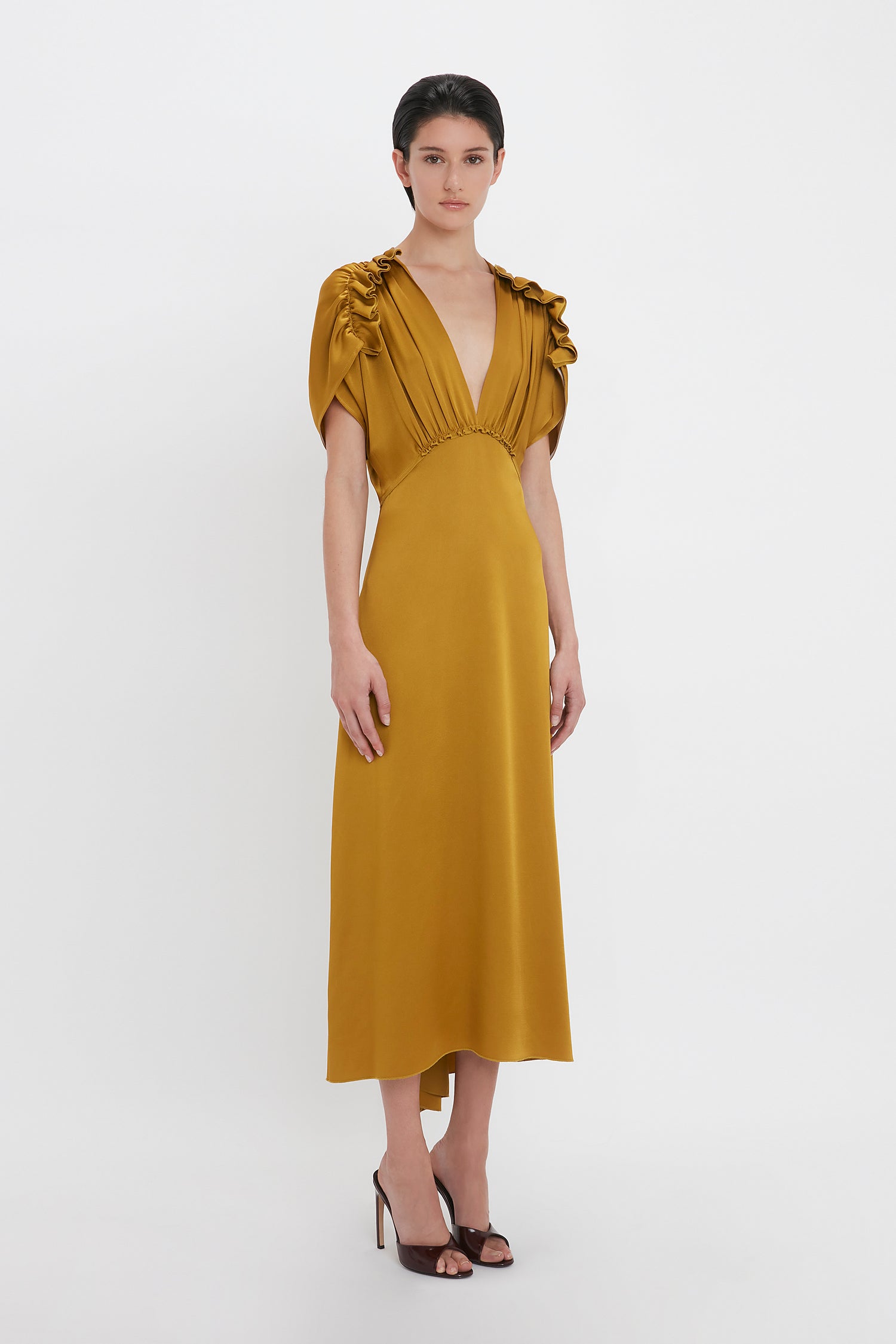 A woman stands in a studio wearing a V-Neck Ruffle Midi Dress In Harvest Gold by Victoria Beckham, paired with black peep-toe high heels. She has short, dark hair and a neutral expression.
