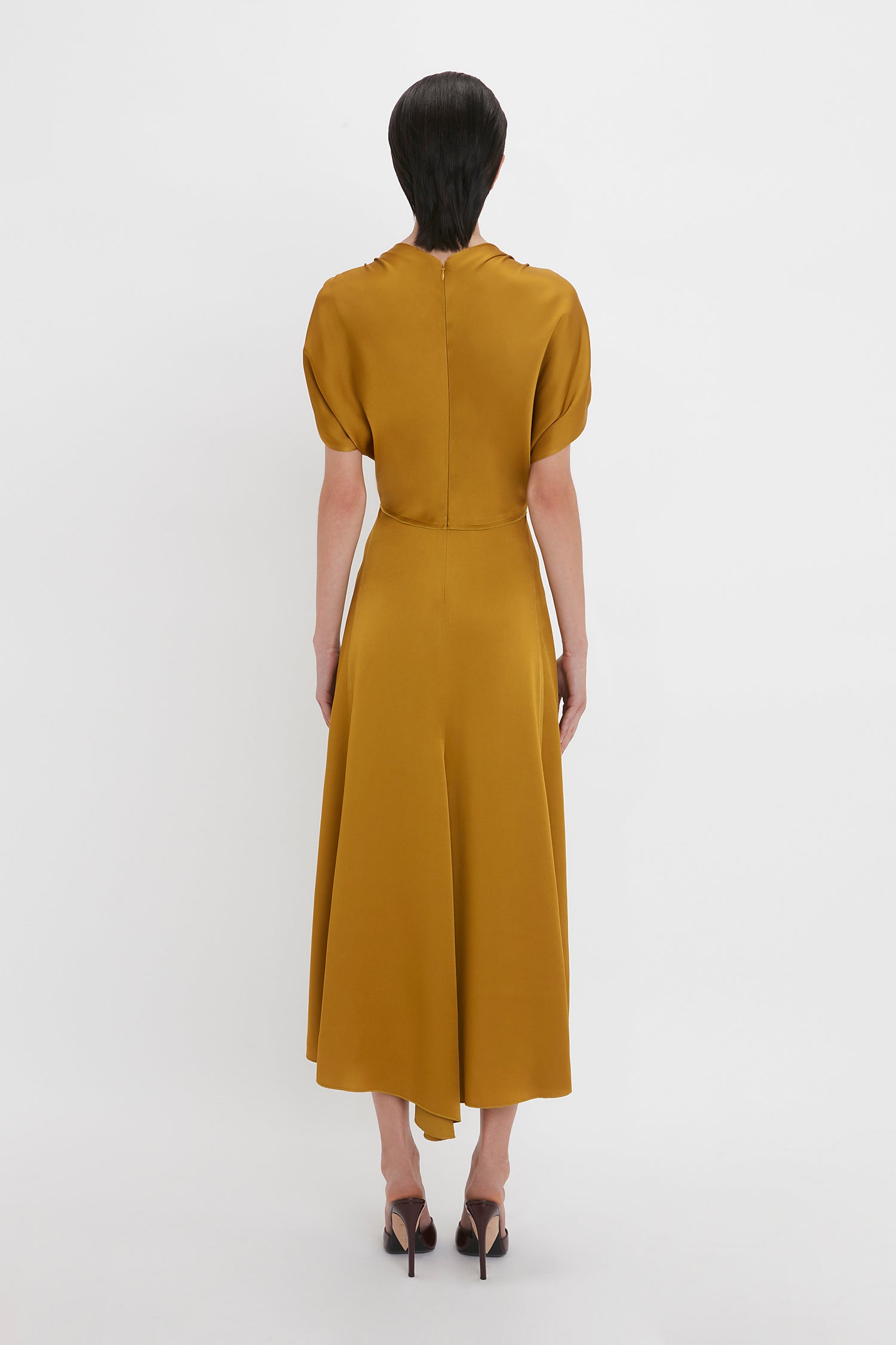 A person with dark hair is standing and facing away, wearing a Victoria Beckham V-Neck Ruffle Midi Dress In Harvest Gold with shoulder frills and black high-heeled shoes against a plain white background.