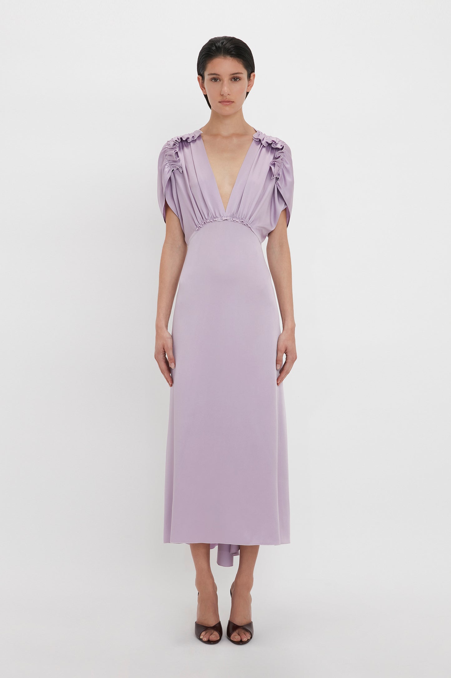 A person stands against a white background, wearing an elegant light purple dress with short, gathered sleeves and a deep V-neck. The ankle-length dress, a V-Neck Ruffle Midi Dress In Petunia from Victoria Beckham, is paired with black heeled shoes.