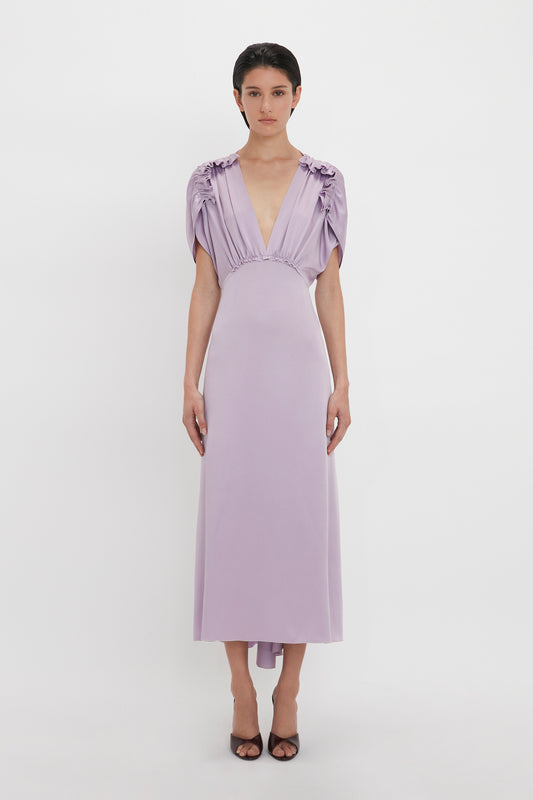 A person stands against a white background, wearing an elegant light purple dress with short, gathered sleeves and a deep V-neck. The ankle-length dress, a V-Neck Ruffle Midi Dress In Petunia from Victoria Beckham, is paired with black heeled shoes.