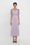 A person stands against a white background, wearing an elegant light purple dress with short, gathered sleeves and a deep V-neck. The ankle-length dress, a V-Neck Ruffle Midi Dress In Petunia from Victoria Beckham, is paired with black heeled shoes.