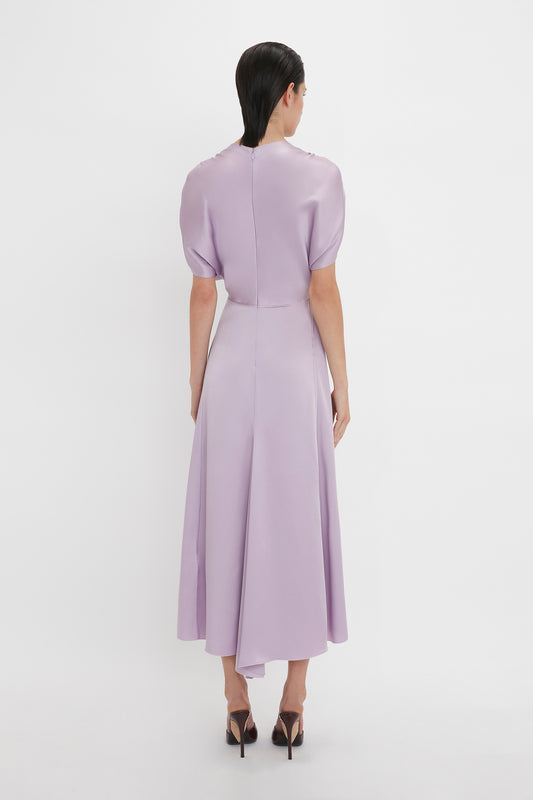 A person stands facing away, wearing a light purple, short-sleeved, mid-length dress with a high neckline and black high heels on a plain white background. This elegant V-Neck Ruffle Midi Dress In Petunia exudes timeless charm reminiscent of the Victoria Beckham brand.