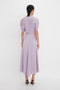 A person stands facing away, wearing a light purple, short-sleeved, mid-length dress with a high neckline and black high heels on a plain white background. This elegant V-Neck Ruffle Midi Dress In Petunia exudes timeless charm reminiscent of the Victoria Beckham brand.