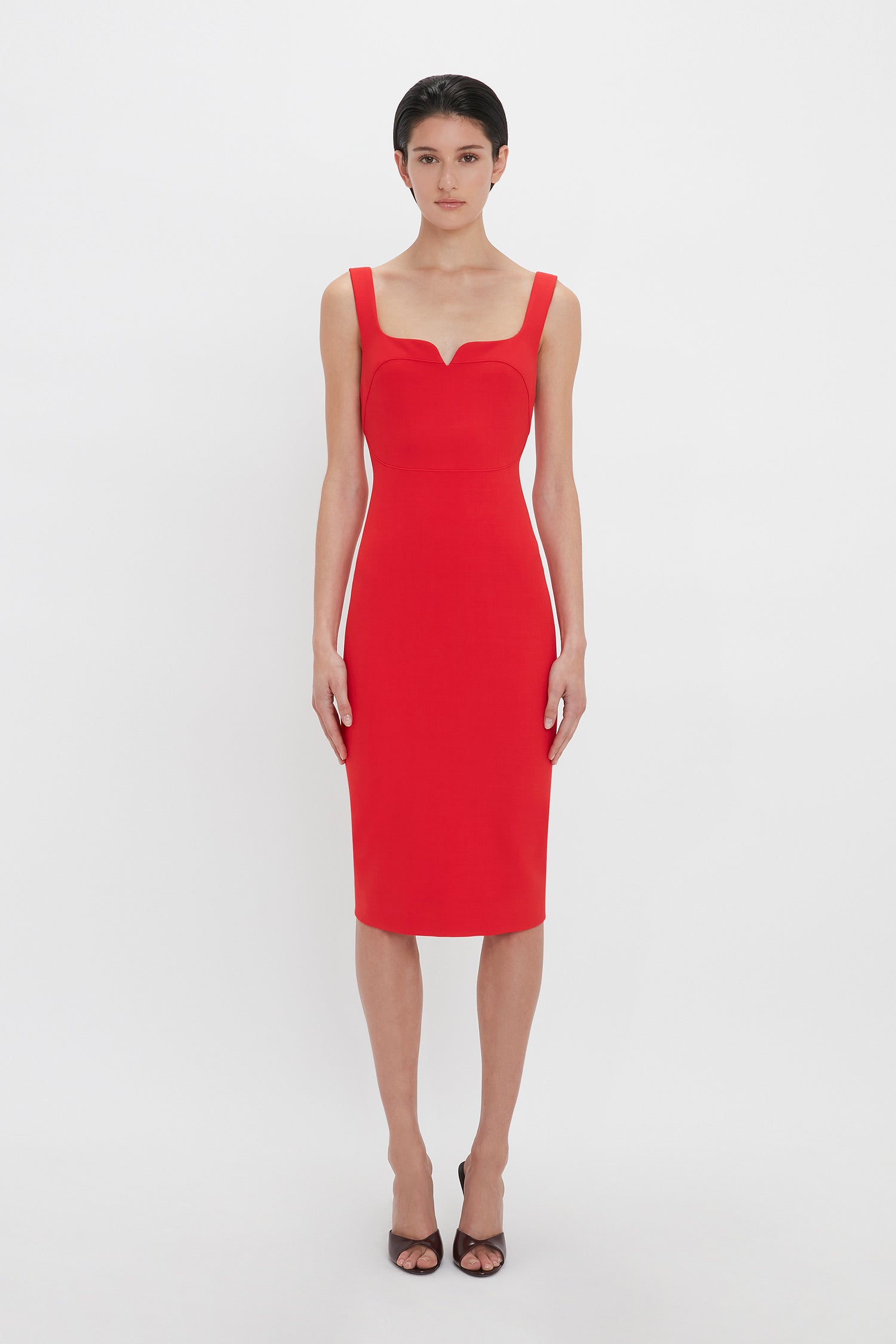 Person standing against a plain background, wearing a Sleeveless Fitted T-Shirt Dress In Bright Red by Victoria Beckham, and black high-heeled shoes.
