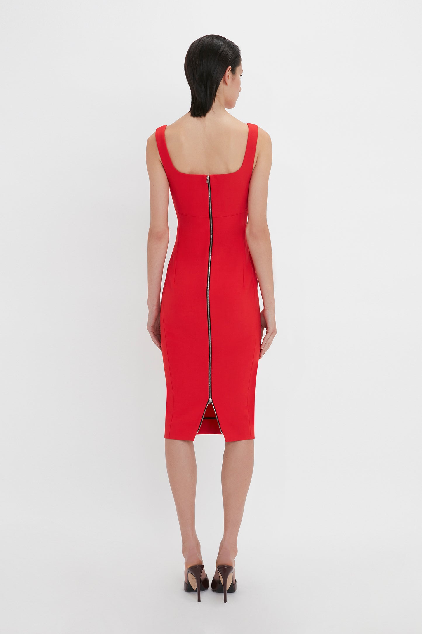 A person in a Sleeveless Fitted T-Shirt Dress In Bright Red by Victoria Beckham standing with their back to the camera, showcasing a visible zipper running from the neckline to the knee-length hem. Made from double wool crepe, the dress has wide shoulder straps.