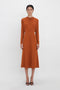 Person wearing a long-sleeved, rust-colored Dolman Midi Dress In Russet by Victoria Beckham with a fitted waist and voluminous underarm drape, standing against a plain white background.