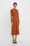 A person with short hair and a neutral expression is wearing a burnt orange **Dolman Midi Dress In Russet by Victoria Beckham** featuring voluminous underarm drape and black open-toed high-heeled shoes. They are standing against a plain white background.