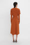 A person stands facing away from the camera, elegantly clad in a Victoria Beckham Dolman Midi Dress In Russet crafted from fluid cady fabric. The rust-colored, knee-length dress features a voluminous underarm drape and is paired with black high-heeled shoes against a white background.