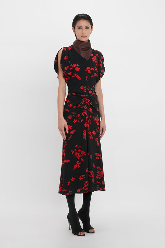 A person stands against a white background wearing a Gathered Waist Midi Dress In Sci-Fi Black Floral by Victoria Beckham, featuring a fit-and-flare silhouette and gathered waist. They accessorize with a brown neck scarf, black open-toe heels, and black stockings.
