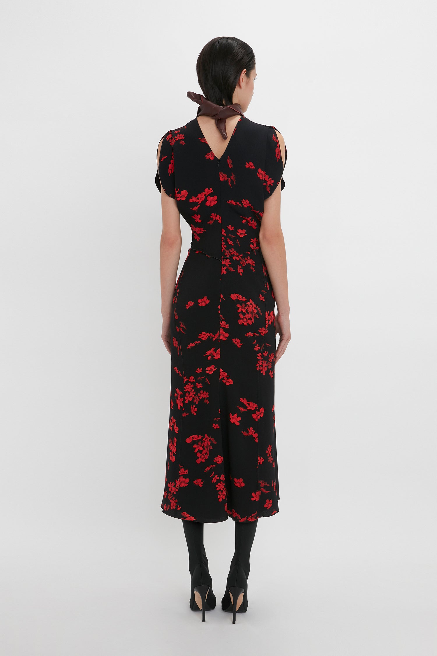 A person with dark hair is standing with their back facing the camera, wearing a Victoria Beckham Gathered Waist Midi Dress In Sci-Fi Black Floral, black stockings, and black high-heeled shoes.