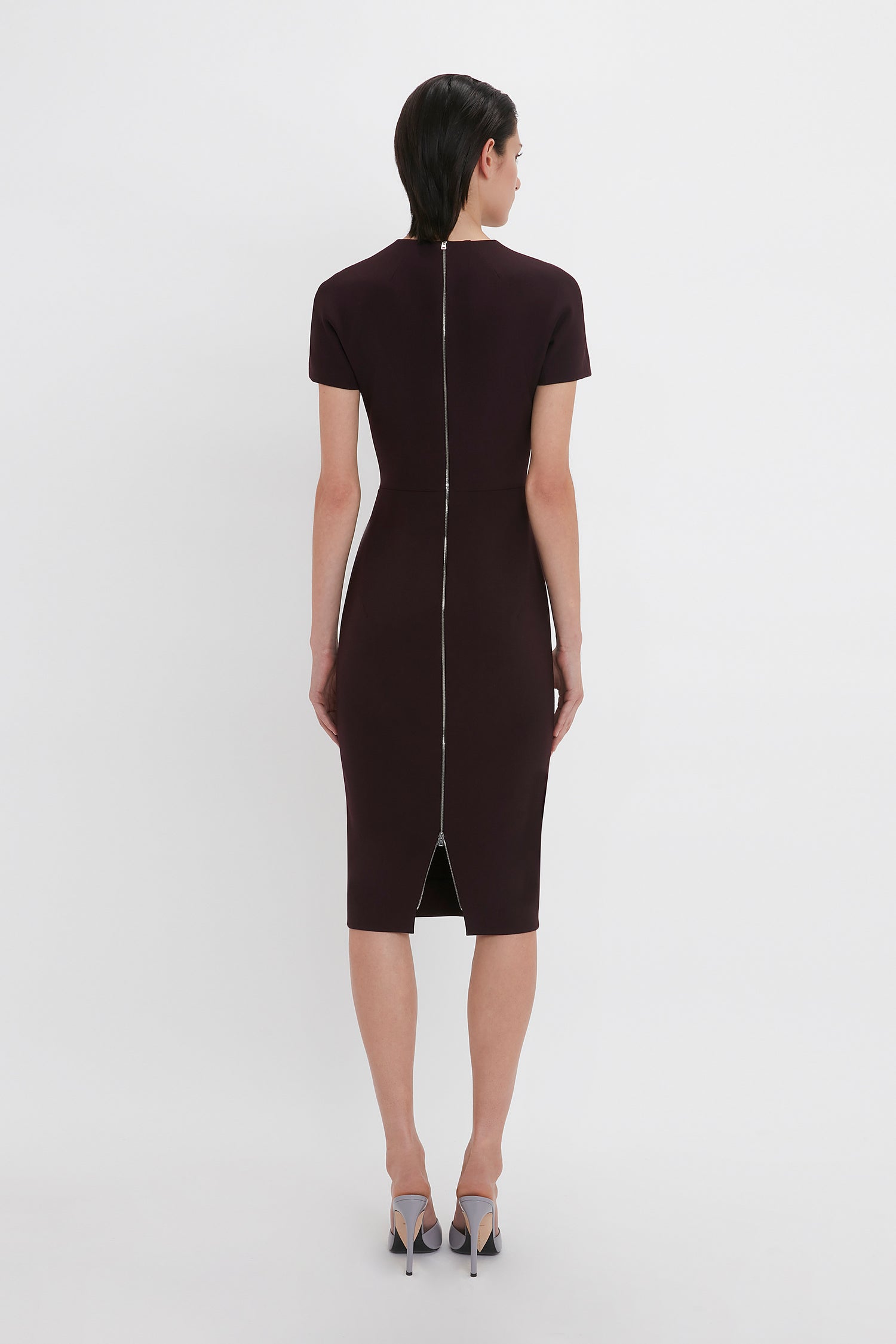 A person with short black hair is seen from the back, wearing a knee-length, short-sleeve Victoria Beckham Fitted T-Shirt Dress In Deep Mahogany with a visible zipper down the middle, and high-heeled shoes.