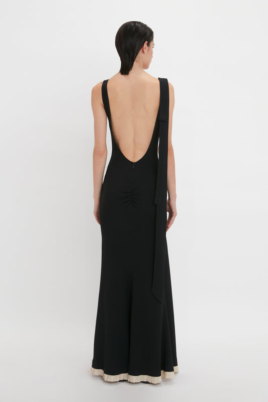 A person wearing the V-Neck Gathered Waist Floor-Length Gown In Black by Victoria Beckham, with wide straps and a floor-length hem, faces away from the camera against a plain white background.