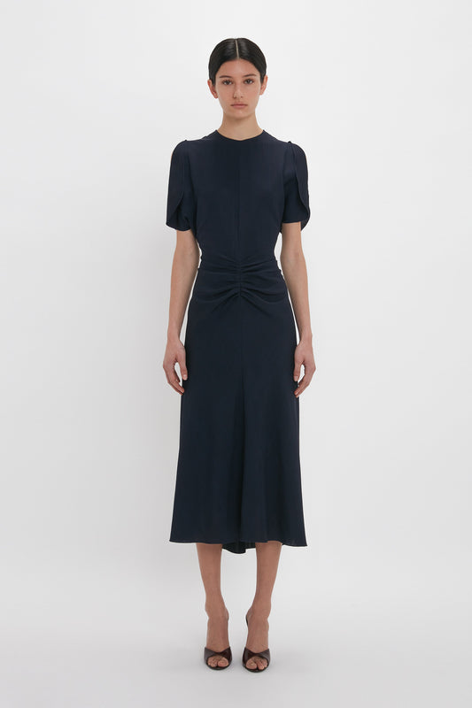 A woman in a navy blue, Gathered V-Neck Midi Dress In Midnight by Victoria Beckham with tulip sleeve detail stands facing the camera. She is wearing brown open-toe heels. The background is plain white.