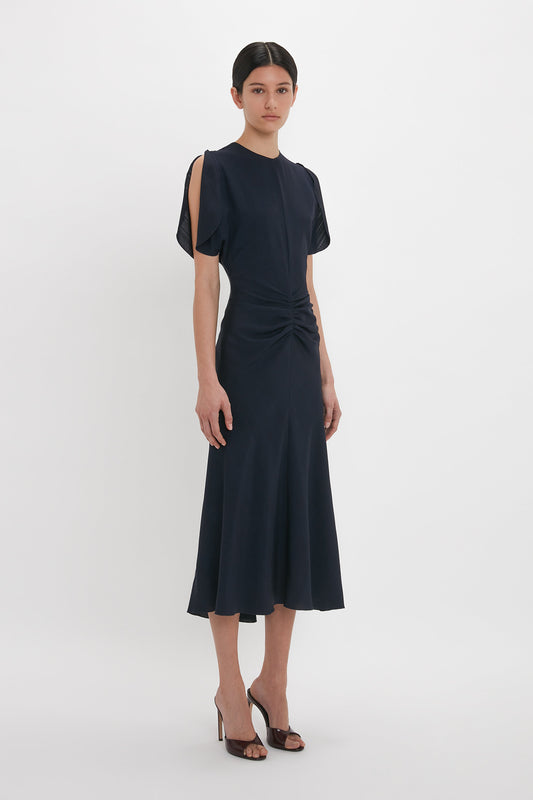 A person stands posing in a dark, short-sleeved, knee-length dress with cut-out shoulders. The Victoria Beckham Gathered V-Neck Midi Dress In Midnight features a fit-and-flare silhouette with a fitted waist and flared skirt. They are wearing high-heeled sandals.