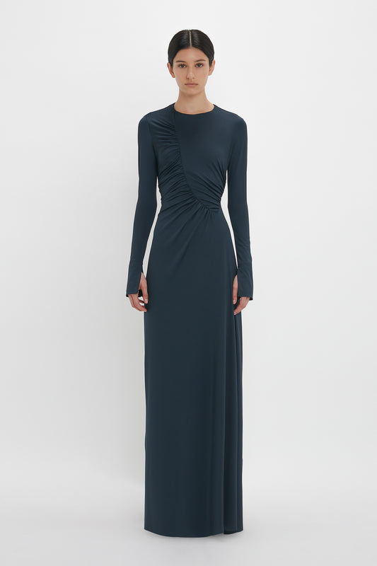 Person standing in a full-length dark blue Ruched Detail Floor-Length Gown In Midnight by Victoria Beckham with long sleeves, embodying understated glamour against a plain white background.