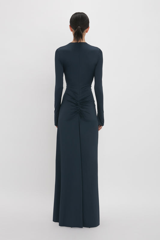 A person stands facing away, wearing a Victoria Beckham Ruched Detail Floor-Length Gown In Midnight: a long-sleeved, floor-length navy dress with ruched detailing at the lower back, exuding understated glamour.