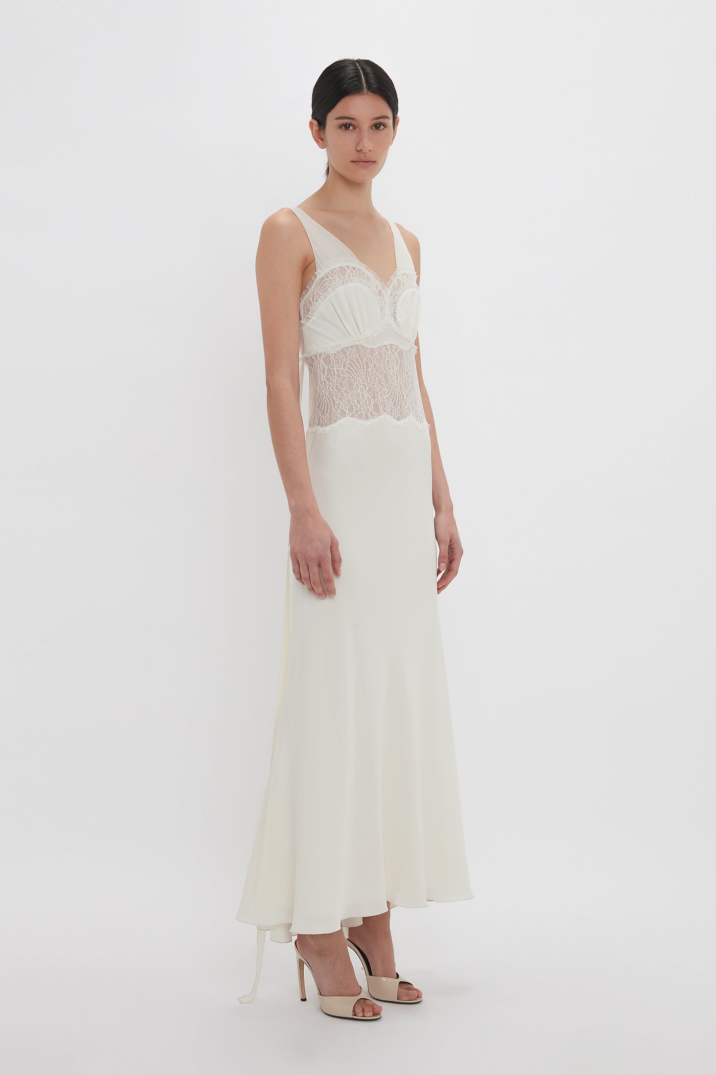 A person in an elegant Ruffle Detail Midi Dress In Ivory by Victoria Beckham with a tactile lace bodice and a silk skirt stands against a plain white background. The sleeveless dress features spaghetti straps and a flowy fit, combining simplicity with refined elegance.