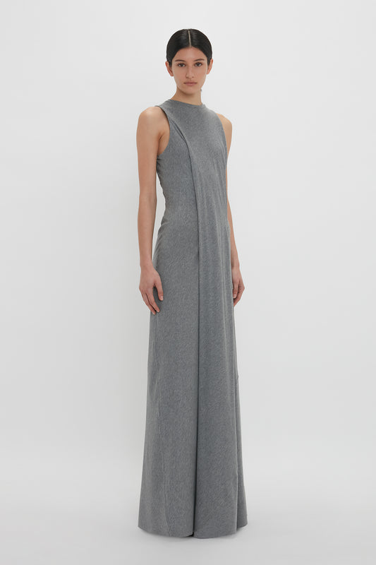 A person stands wearing a sleeveless, floor-length Frame Detailed Maxi Dress In Titanium by Victoria Beckham against a plain white background.