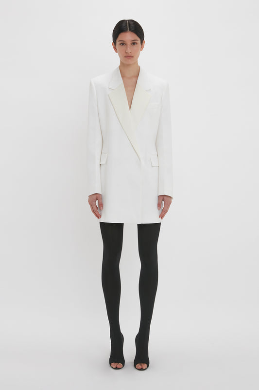 A woman in a Victoria Beckham Exclusive Fold Shoulder Detail Dress In Ivory and black tights stands against a plain white background.