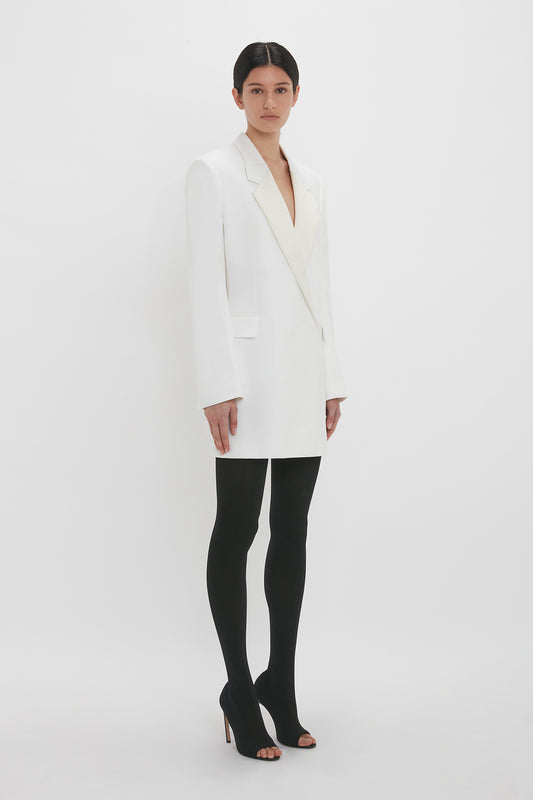 A woman stands in a white studio, wearing a long white double-breasted cut blazer dress with black tights and open-toe heels, looking directly at the camera.
