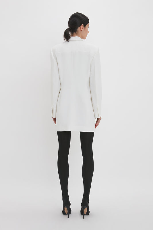 A woman viewed from behind, wearing a Victoria Beckham Exclusive Fold Shoulder Detail Dress In Ivory, paired with black tights and high heels, stands against a white background.