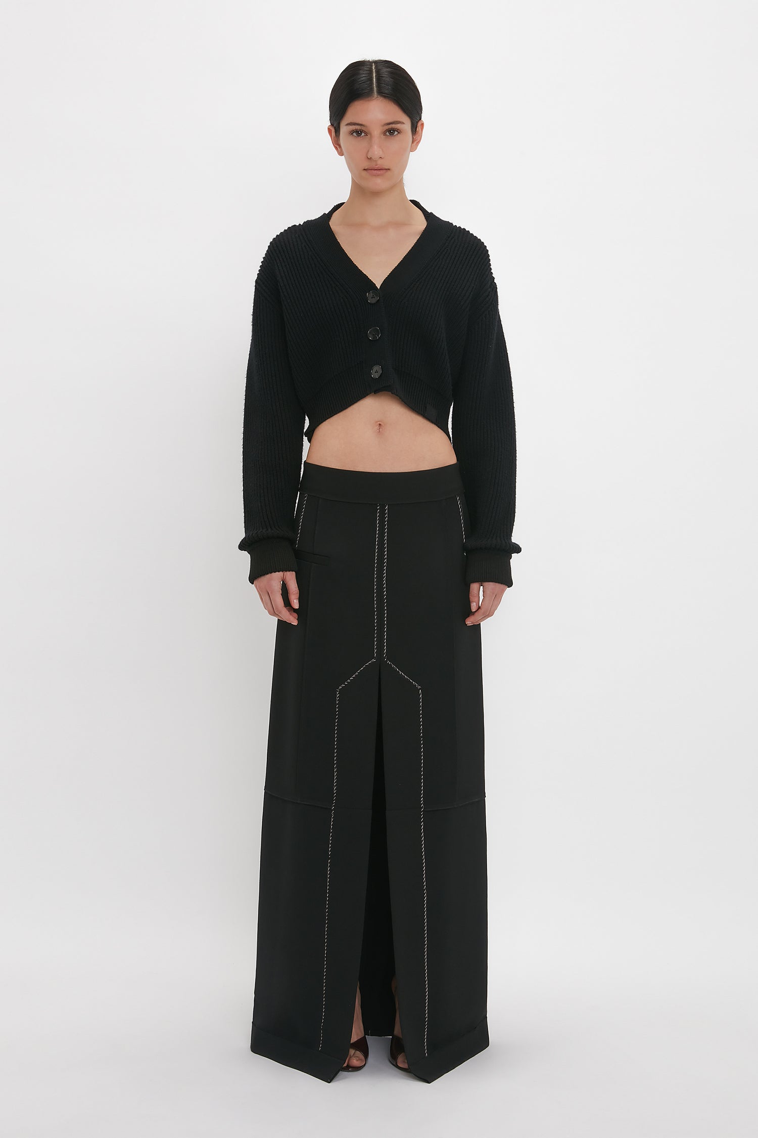 A person stands against a white background, wearing a Cropped V-Neck Cardigan In Black by Victoria Beckham and a long black skirt with stitched details and a front slit.